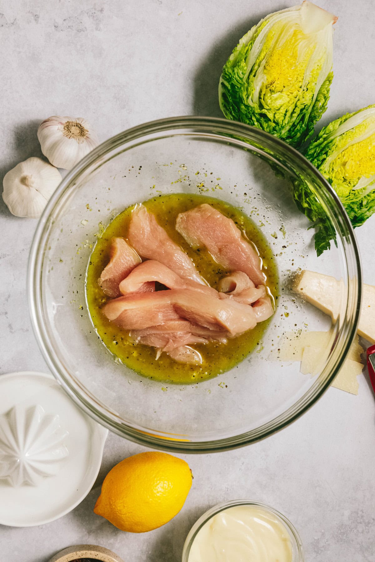 Glass bowl with chicken strips marinating in olive oil and seasoning. Surrounding items include romaine lettuce, garlic bulbs, lemon, mayo, parmesan cheese, and a lemon juicer on a countertop.