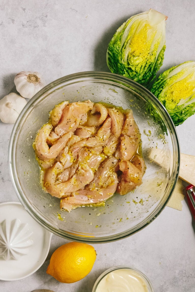 A bowl of marinated chicken strips surrounded by ingredients including garlic, baby romaine lettuce, lemon, mayonnaise, and cheese on a light countertop.