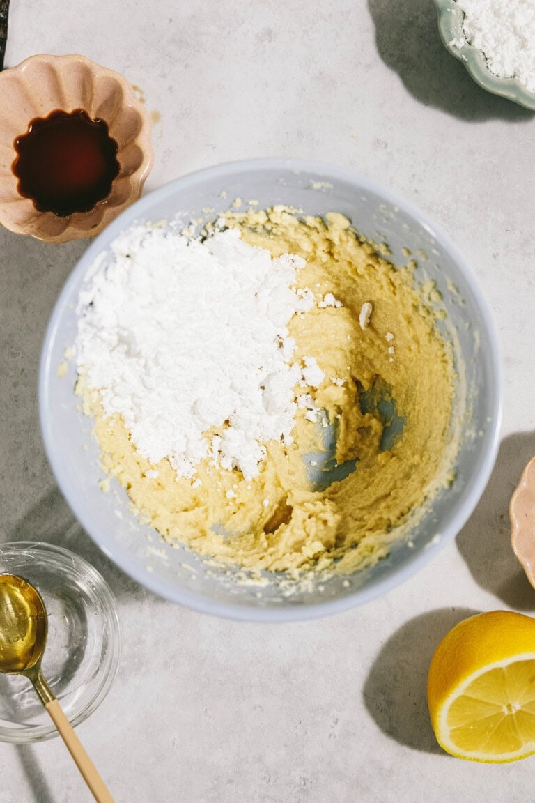 A mixing bowl with yellow batter and white powder sits on a surface surrounded by a lemon, a glass with a honey dipper, and a small bowl of vanilla extract.