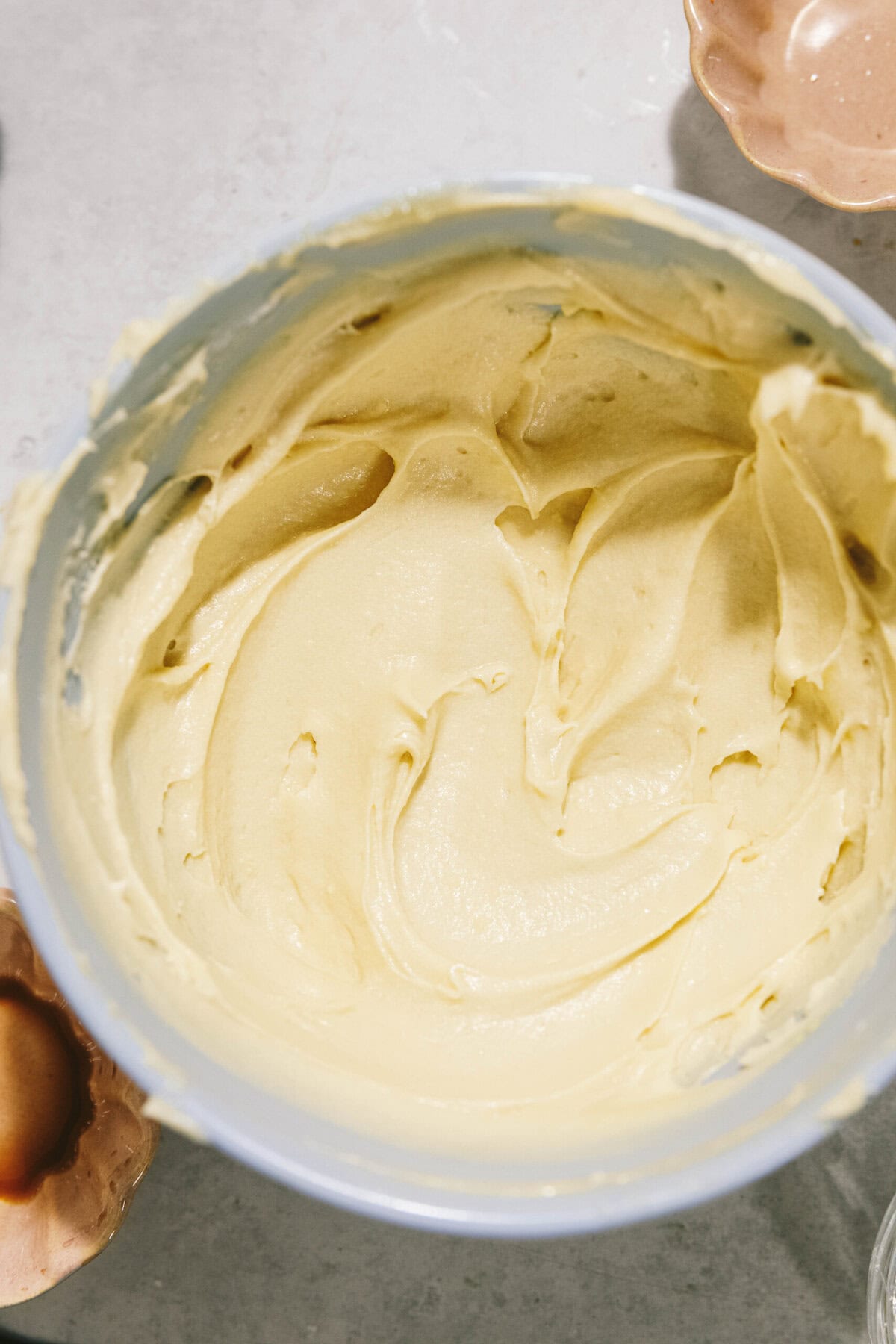 A bowl filled with smooth, creamy batter on a light-colored surface with a few kitchen utensils blurred in the background.