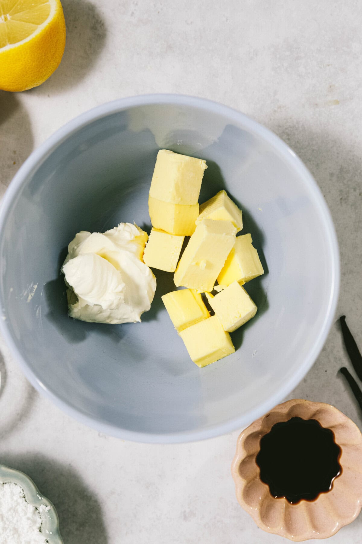 A blue bowl containing several cubes of butter and a dollop of cream cheese sits on a countertop. Nearby are a lemon, a small bowl with a dark liquid, and a dish with a white substance.