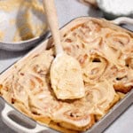 A baking dish of freshly iced cinnamon rolls, with a spatula placed on top. Nearby are a mixing bowl with leftover icing and a small bowl of powdered sugar.