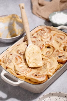 A baking dish of freshly iced cinnamon rolls, with a spatula placed on top. Nearby are a mixing bowl with leftover icing and a small bowl of powdered sugar.