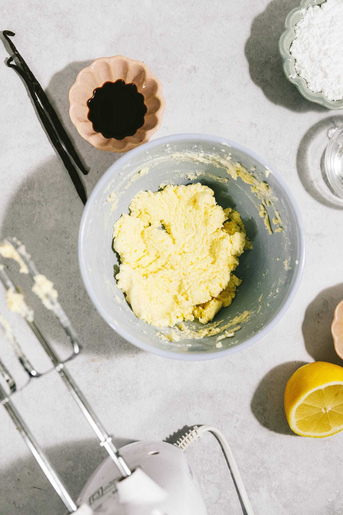 A mixing bowl filled with a yellow dough-like mixture surrounded by vanilla beans, a small bowl with dark liquid, powdered sugar, a lemon, and a hand mixer on a countertop.