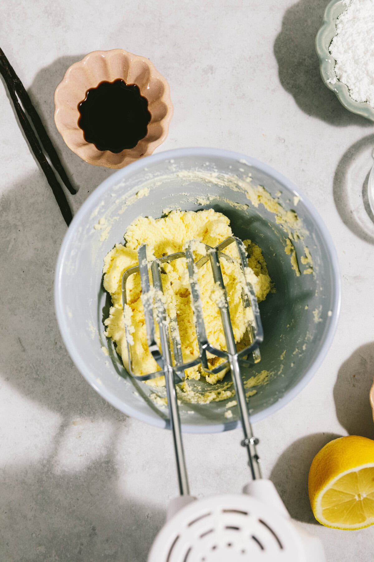 A mixer with yellow batter, a bowl with dark liquid, a lemon, and a bowl with white powder on a white surface.