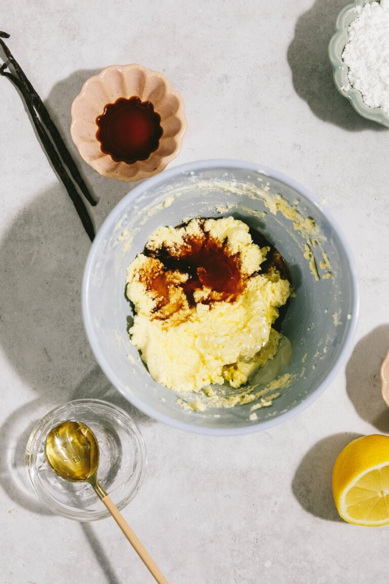 A mixing bowl with creamed butter and vanilla extract, surrounded by ingredients including vanilla beans, a bowl of dark liquid, powdered sugar, lemon, and a glass dish with a golden spoon.