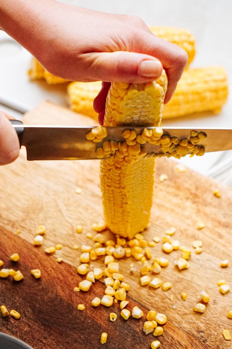 A person is cutting kernels off a cob of corn using a knife on a wooden cutting board.