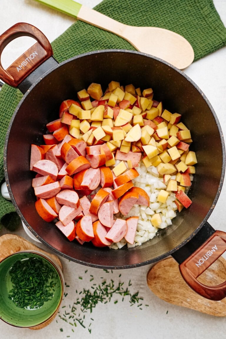 A pot filled with diced potatoes, sliced sausages, and chopped onions placed on a kitchen counter with green towels, a wooden spoon, a small bowl of herbs, and some scattered herbs.