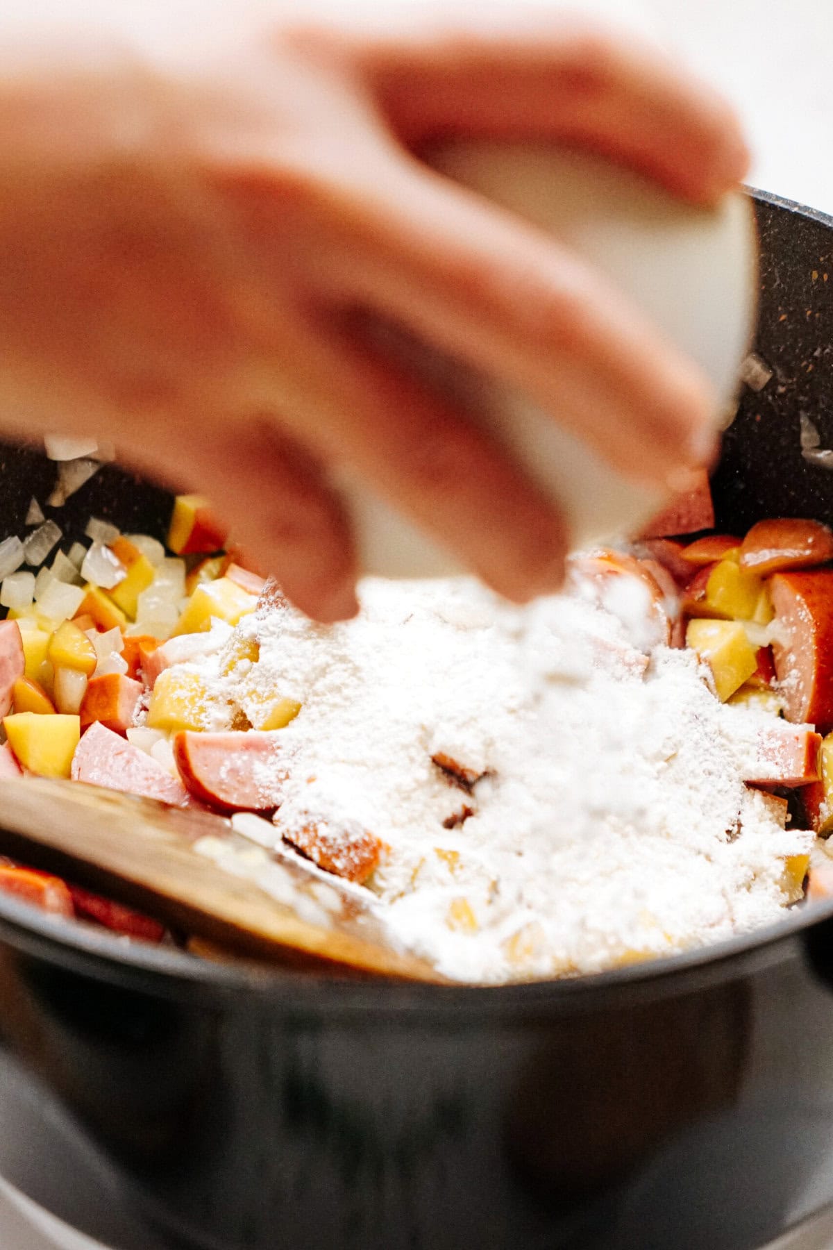 A person adds flour to a mixture of sliced sausages, potatoes, and onions cooking in a black pot.