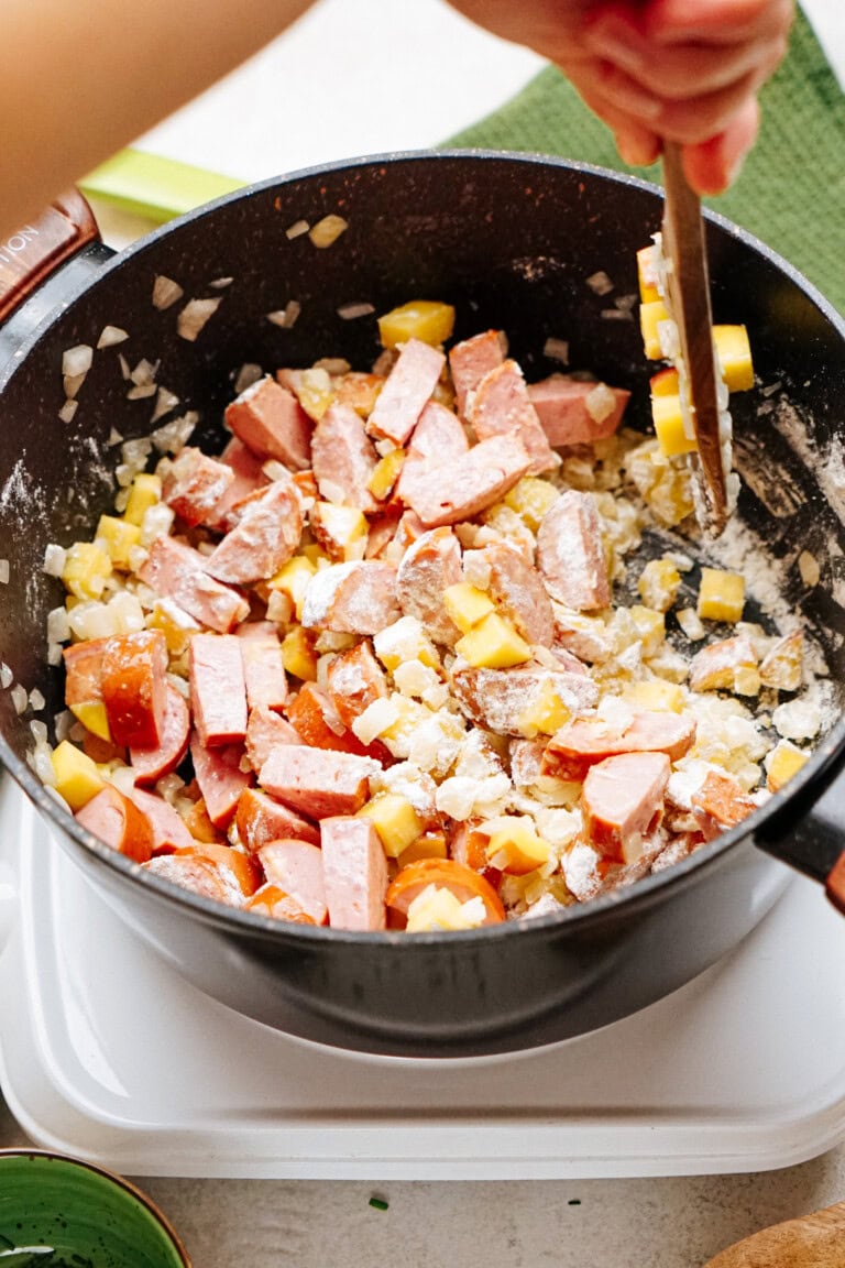 A person stirs a pot containing chopped sausage, diced vegetables, and onions, with some flour apparent in the mix.