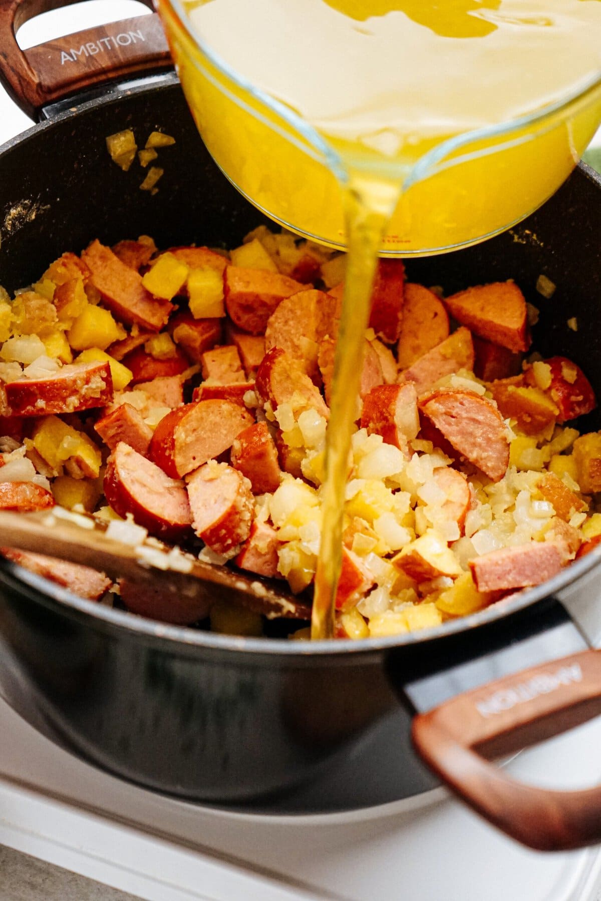 A glass measuring cup pouring broth into a black pot filled with chopped sausages, diced potatoes, and onions, with a wooden spoon inside the pot.