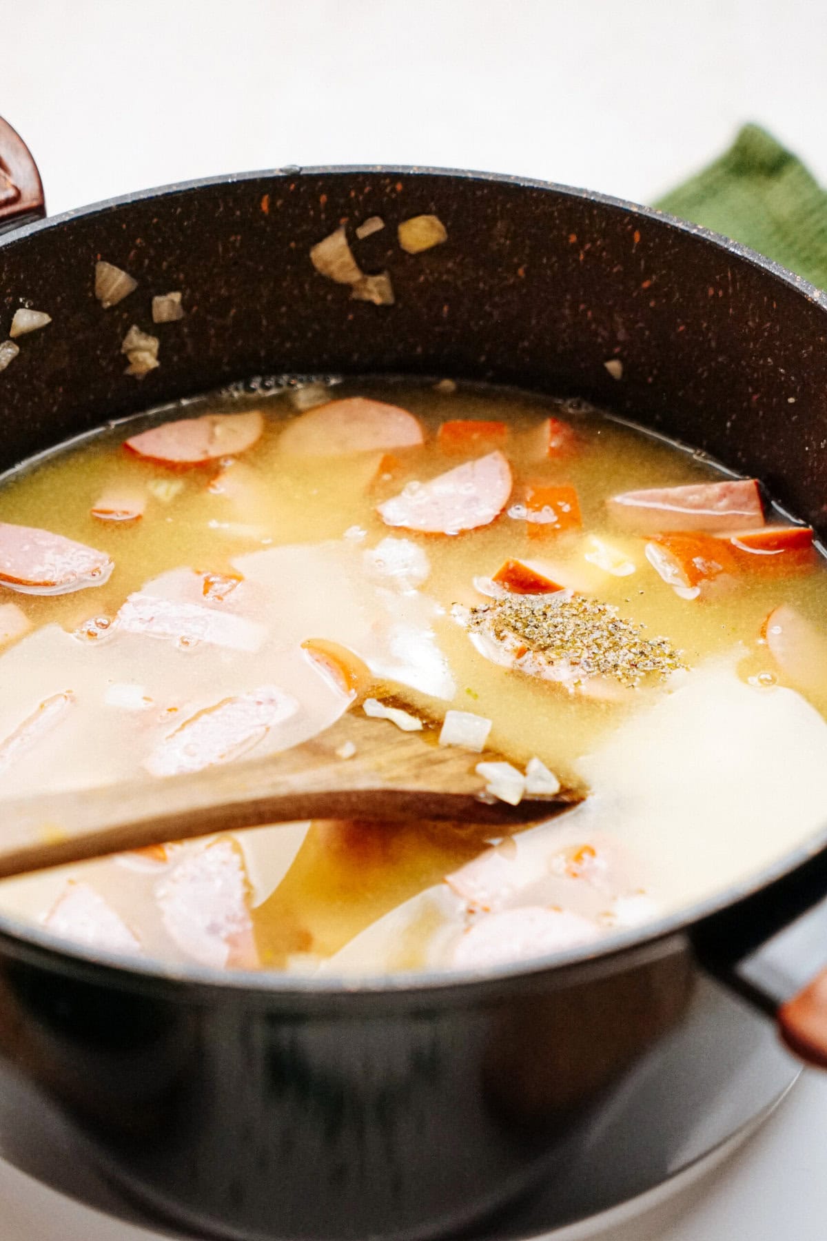 A pot of soup containing sliced sausages, broth, and chopped vegetables with a wooden spoon.