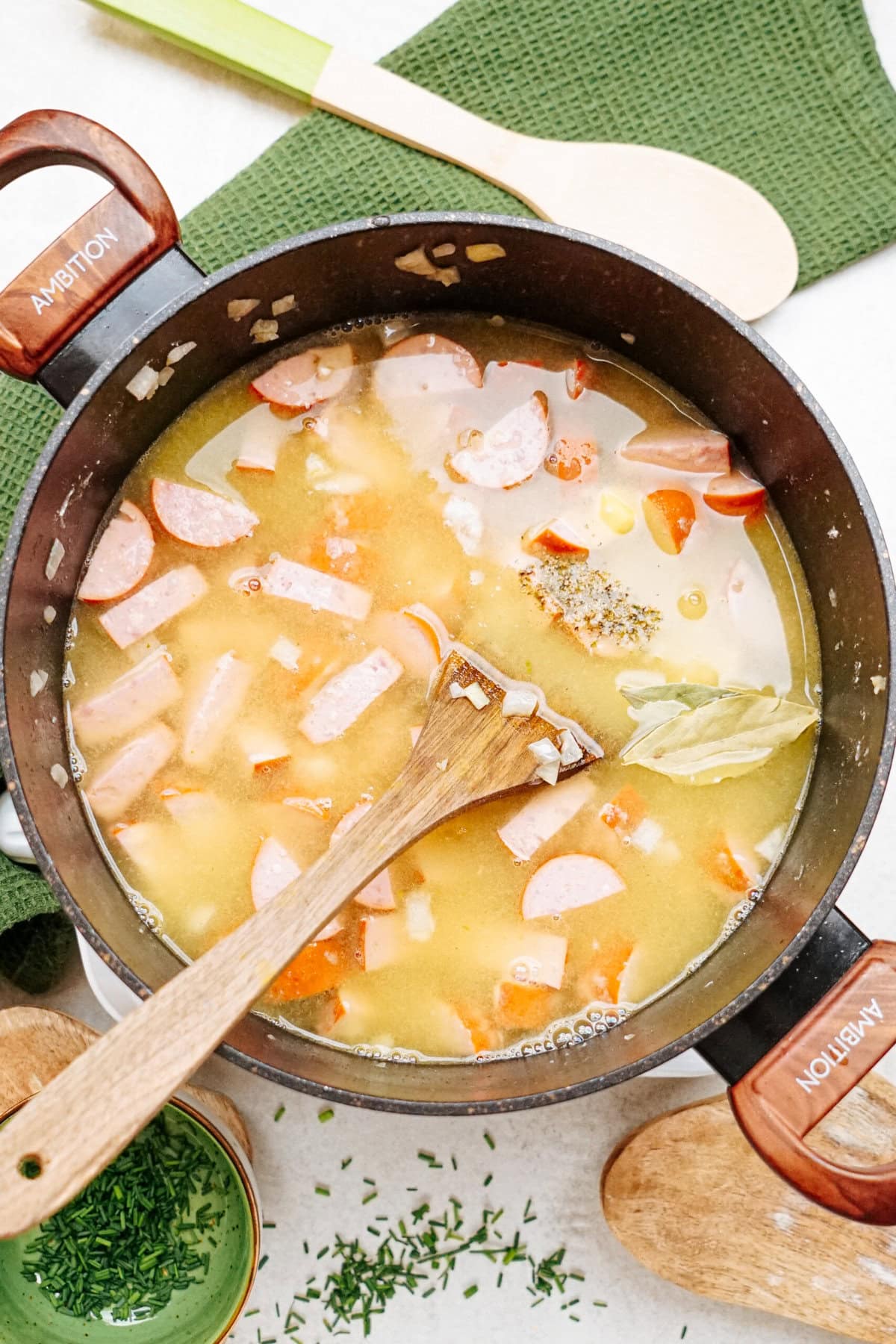 A pot of soup with sausages, sliced carrots, potatoes, and broth. A wooden spoon rests in the pot while ingredients are visible around the pot, including a bay leaf floating on top.