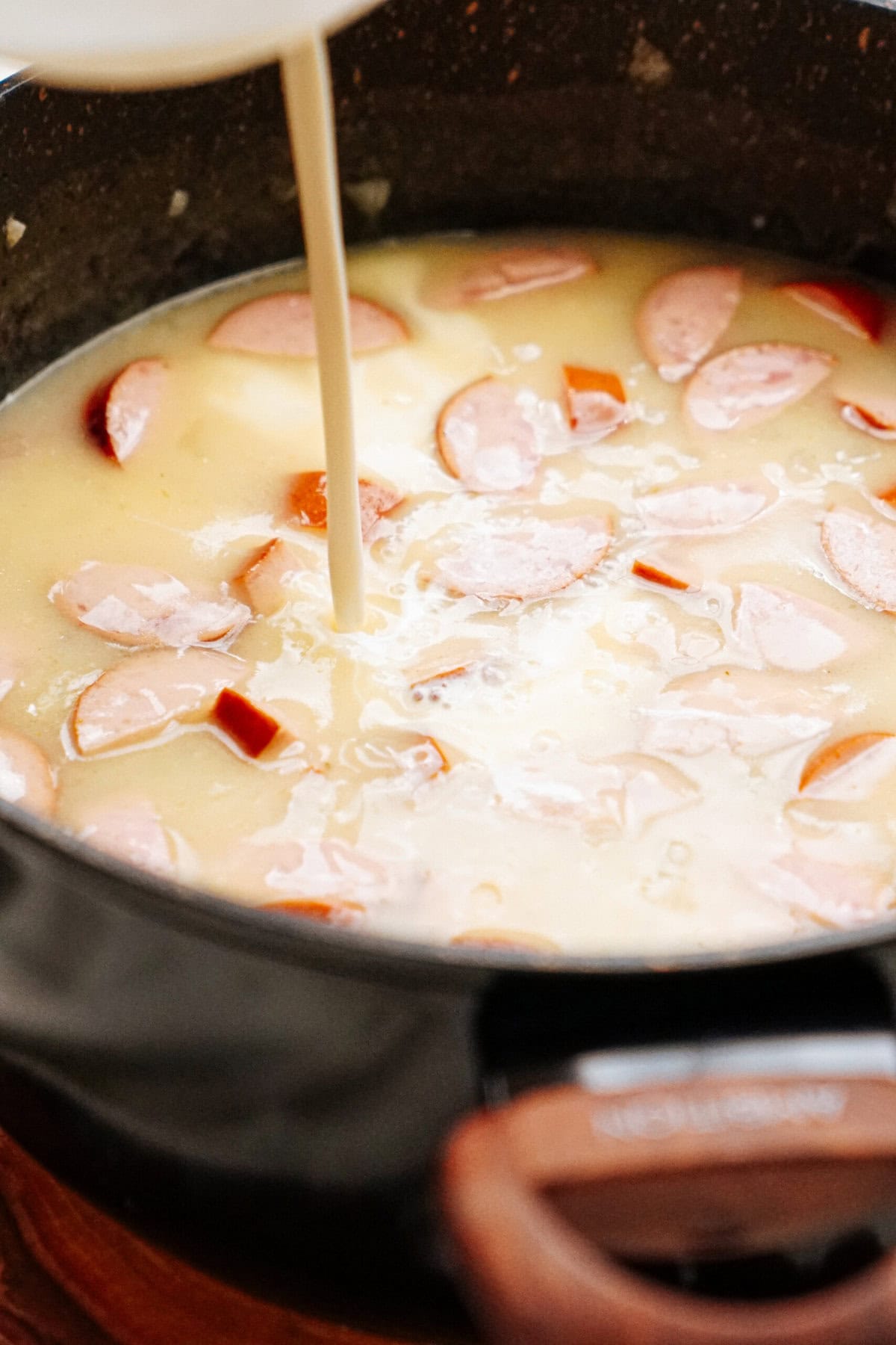 Close-up of sliced sausages in a creamy, pale soup being poured into a dark pot. The mixture appears chunky with visible meat pieces.