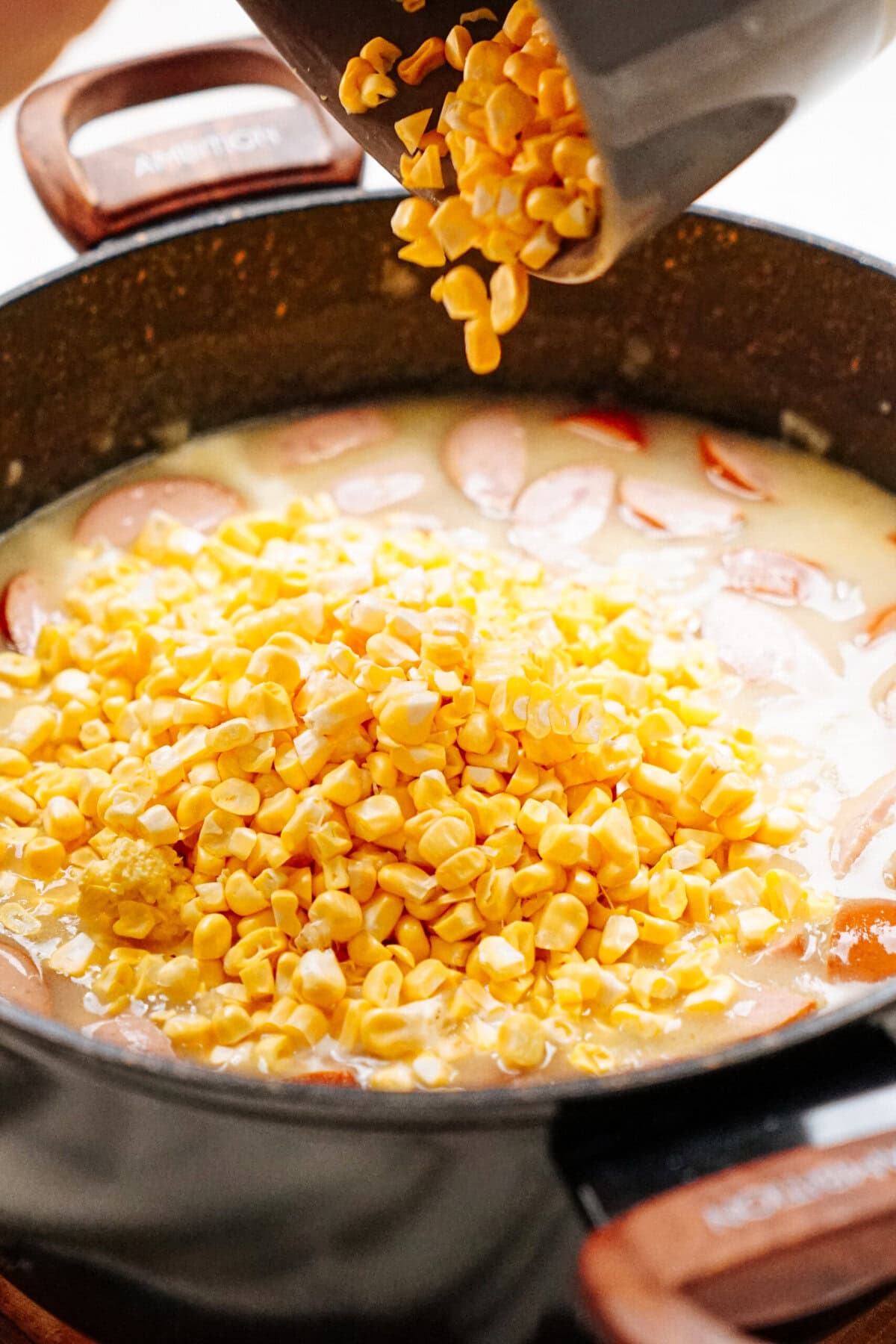 Corn kernels being poured into a pan containing sliced sausages and sauce.