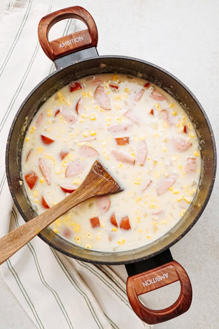 A pot of creamy soup with sliced sausages, corn, and diced vegetables, stirred with a wooden spoon, on a white and green striped kitchen towel.