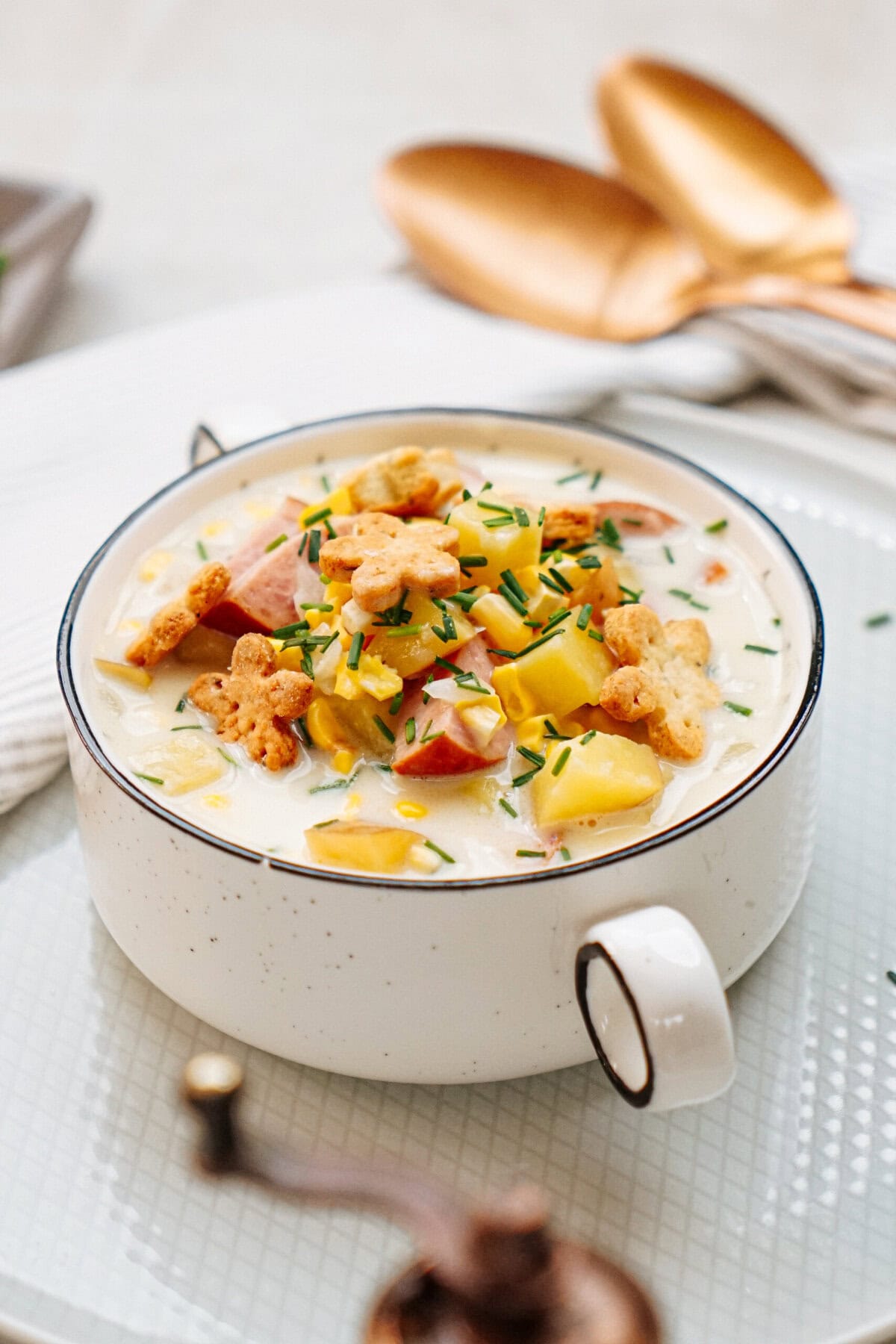 A white ceramic mug filled with creamy sausage corn chowder garnished with diced vegetables, herbs, and cracker toppings. Two golden spoons are placed in the background.