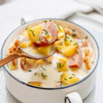 A bowl of creamy sausage corn chowder containing chunks of sausage, potatoes, and garnished with chopped chives. A spoon holding some of the soup is above the bowl.