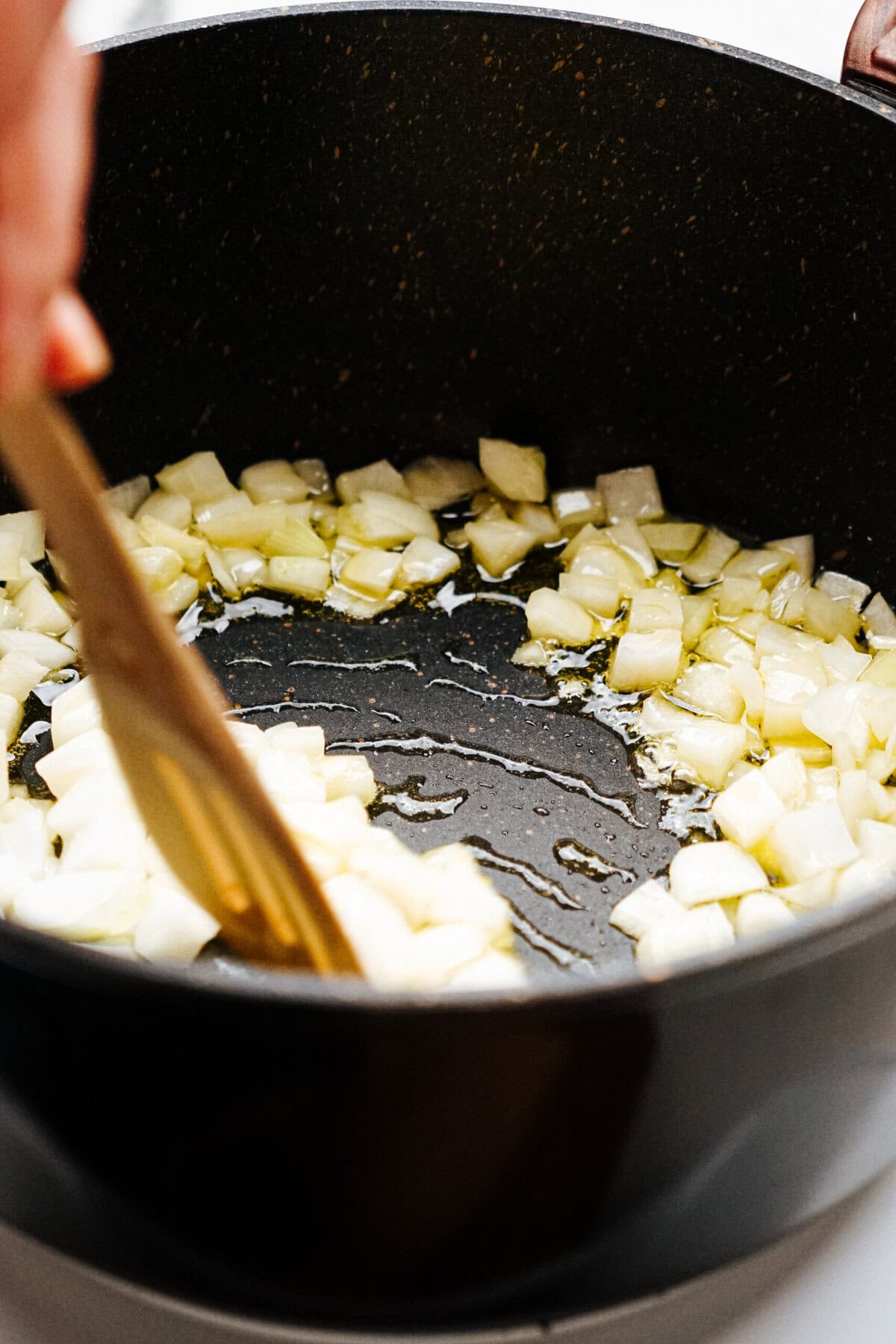 A person stirs chopped onions in a black pot with a wooden spoon.