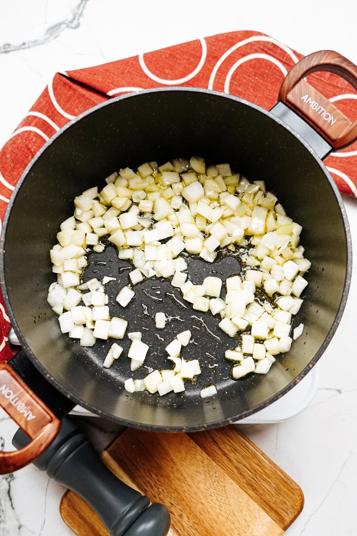 A pan with diced onions sautéing on a stove. A red napkin with a circle pattern is partially visible in the background.