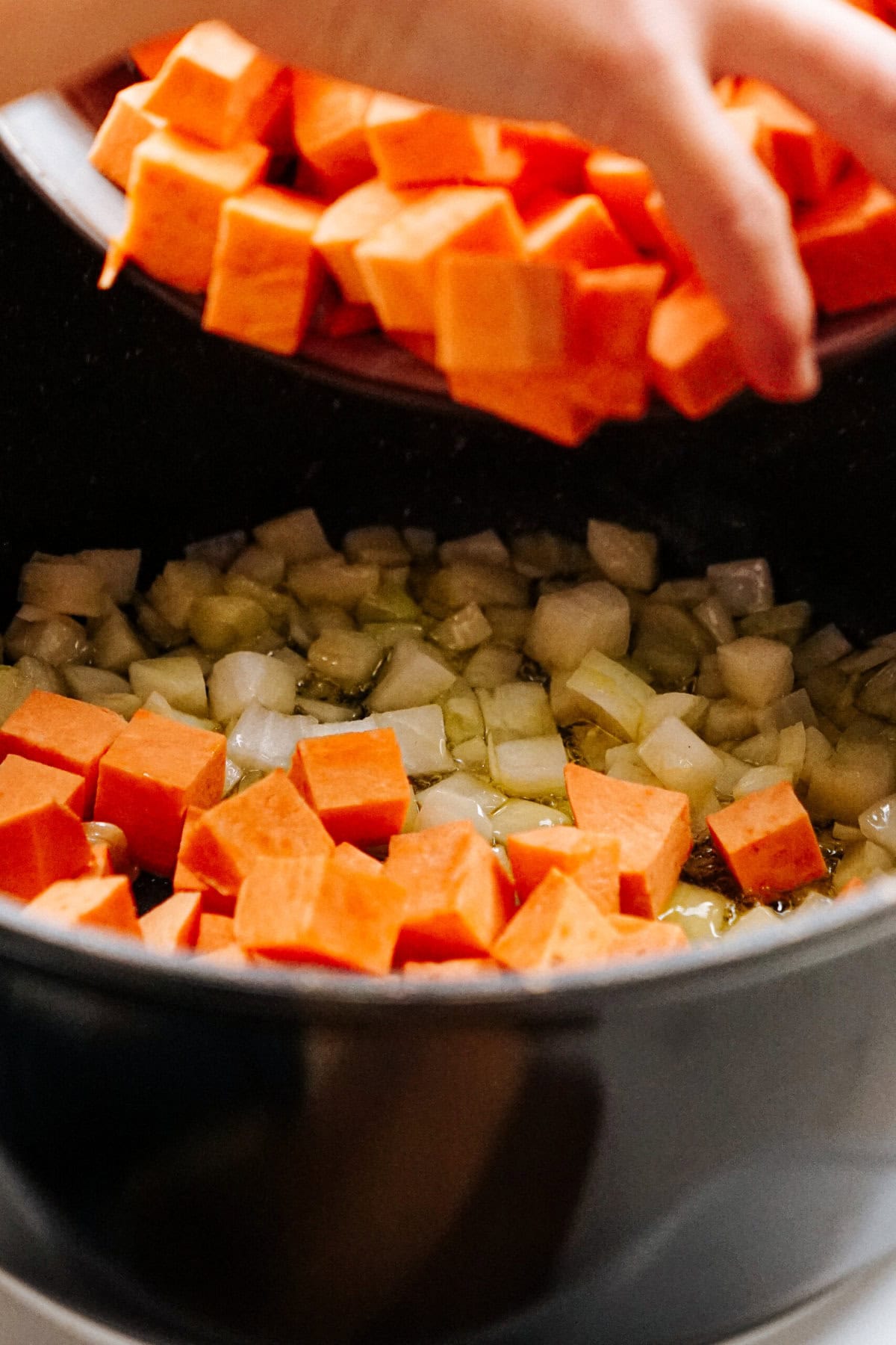 A hand is adding a bowl of cubed sweet potatoes into a black pot containing sautéed onions.