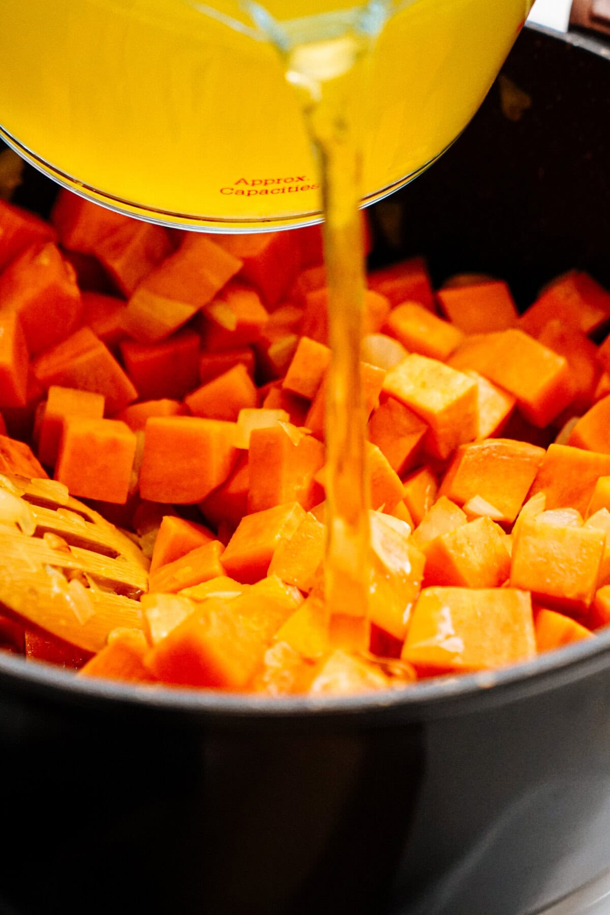 A pot filled with cubed sweet potatoes is shown, with a stream of yellow liquid being poured over them from a measuring cup.