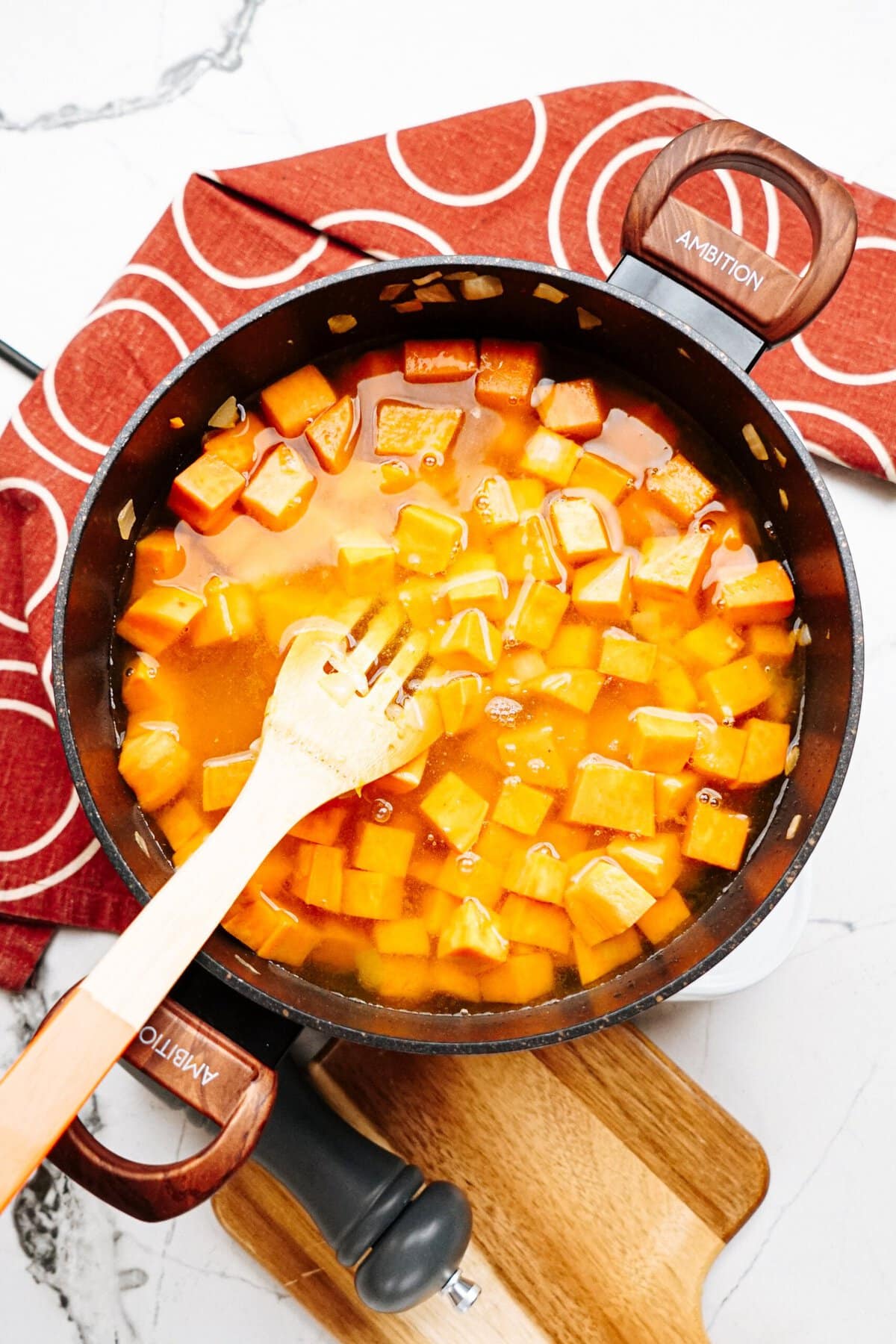 A pan with cubed sweet potatoes being cooked in liquid, stirred with a wooden spatula. A red patterned cloth is beneath the pan on a white countertop. A wooden cutting board is partially visible.