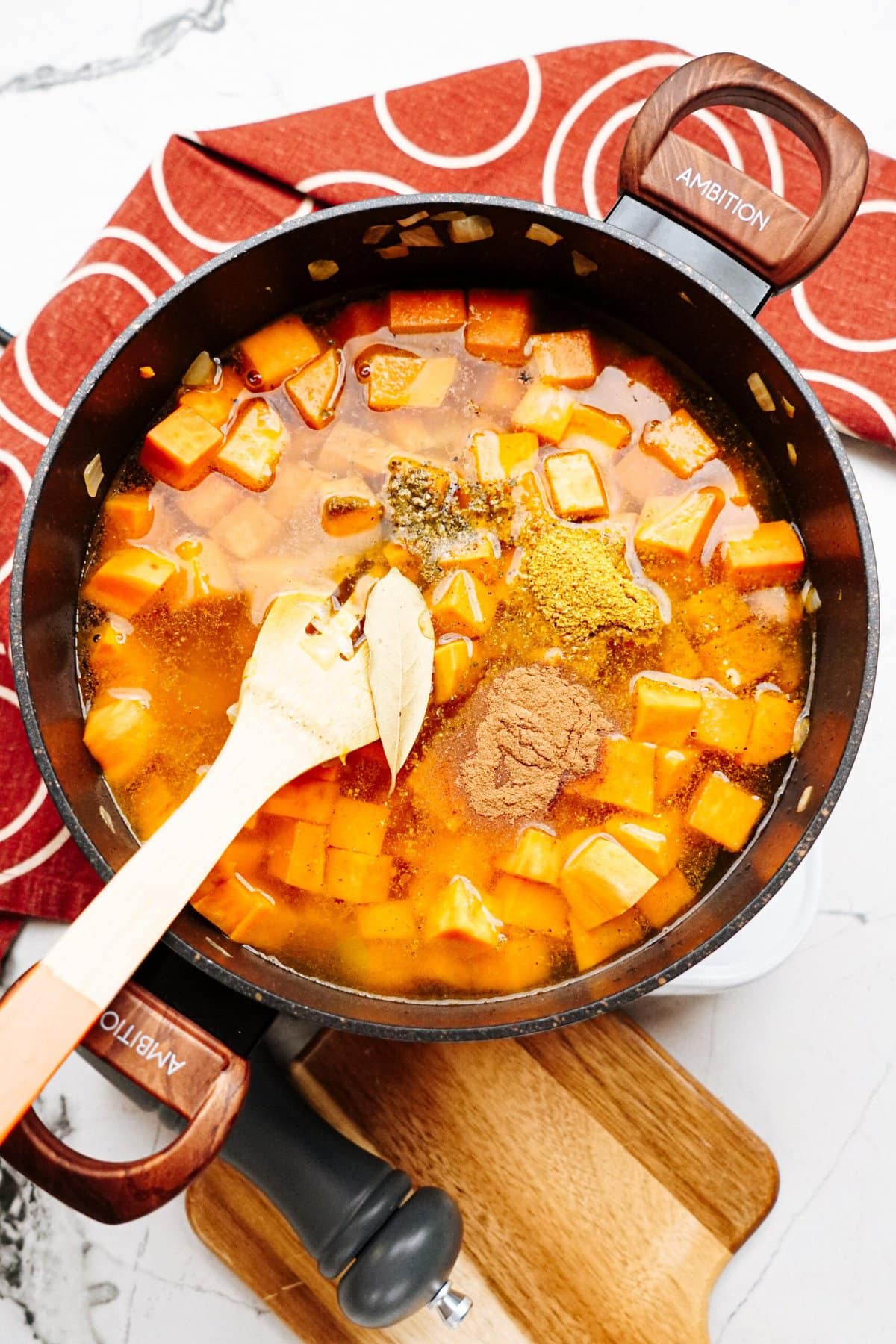 A saucepan with diced sweet potatoes, spices, and broth is being stirred with a wooden spatula. It sits on top of a wooden cutting board with a red cloth napkin in the background.