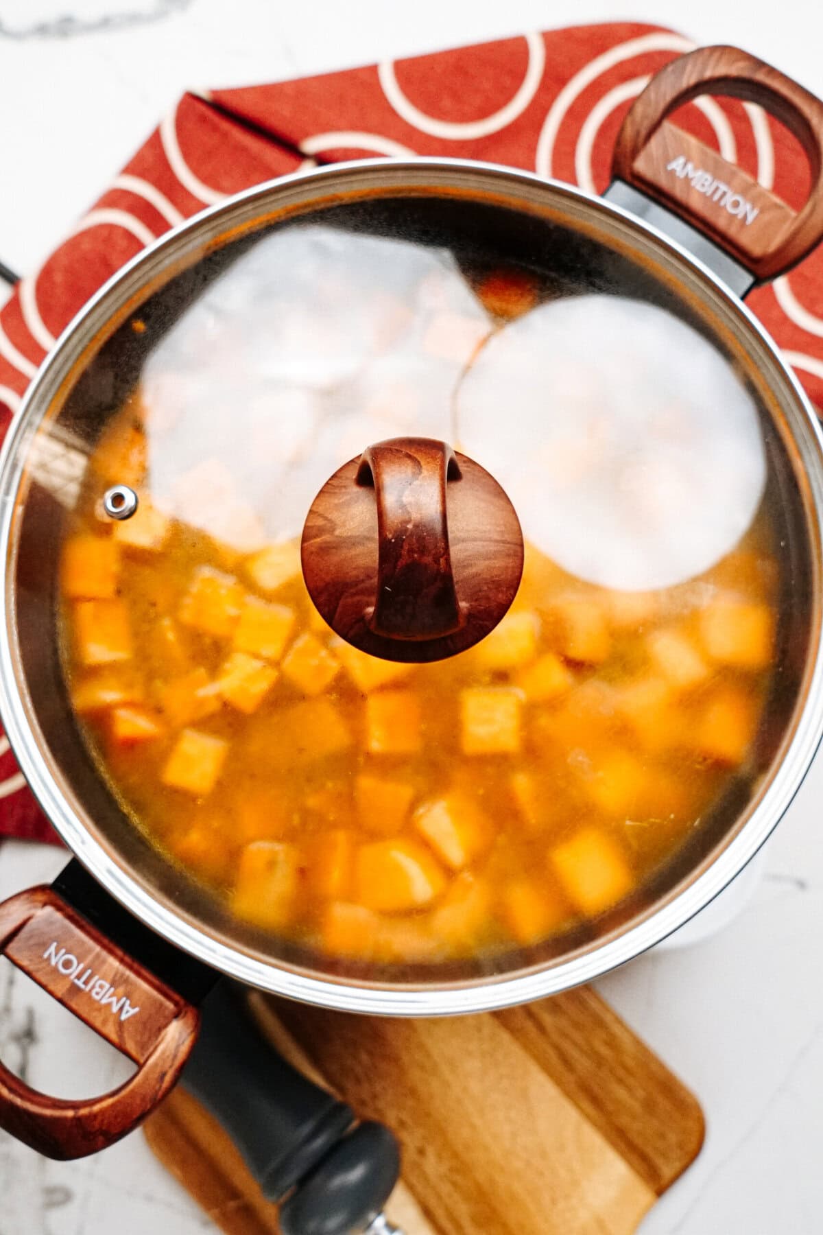 A pot partially covered with a glass lid, showing cubed vegetables cooking in a yellowish broth, sits on a wooden board atop a red cloth with white circular patterns.