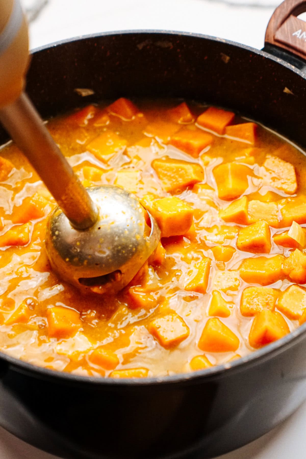 Blender partially submerged in a pot containing chunks of orange vegetables being cooked in a broth.