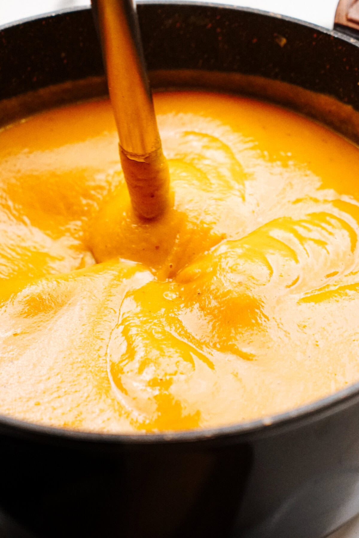 Close-up of an immersion blender blending a pot of creamy orange soup.