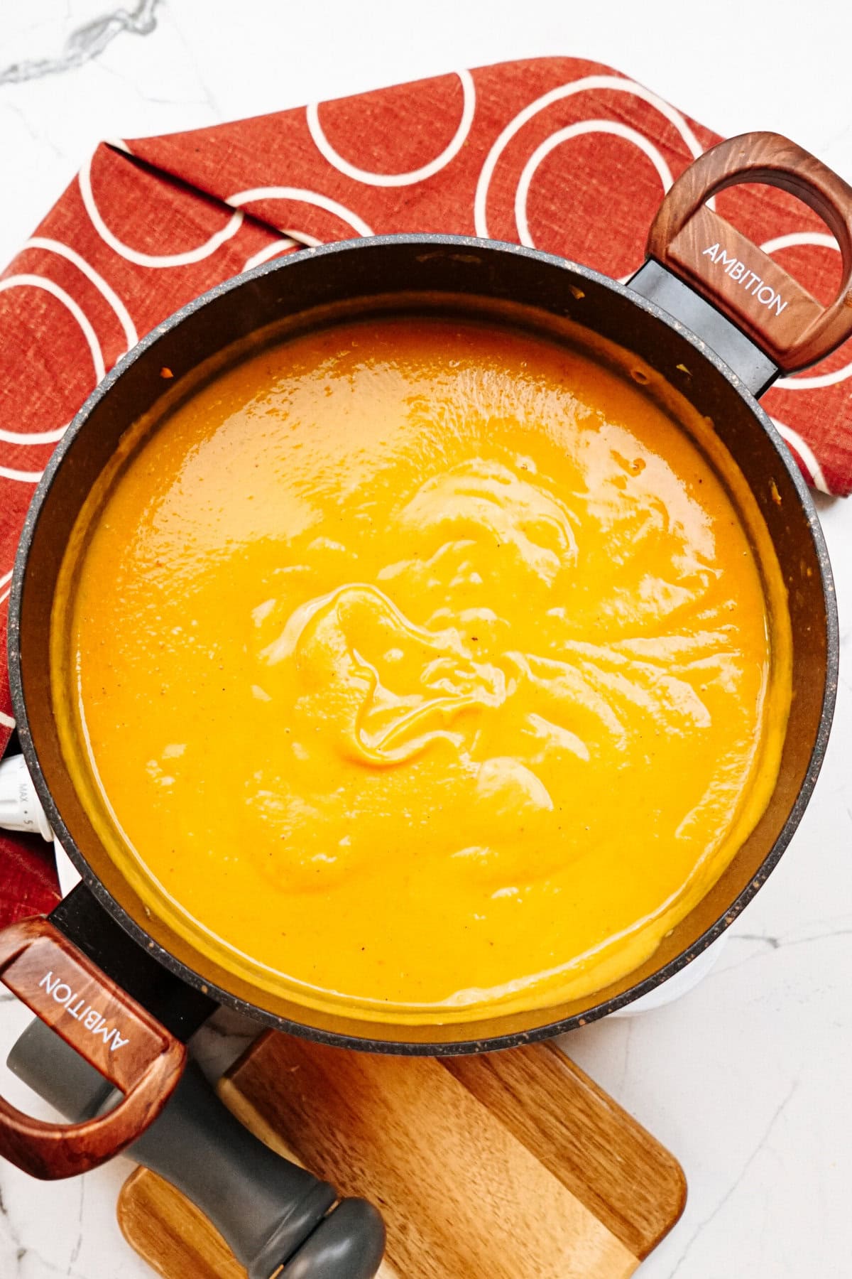 A pot filled with creamy, orange soup sits on a cutting board, with a red and white patterned cloth nearby.