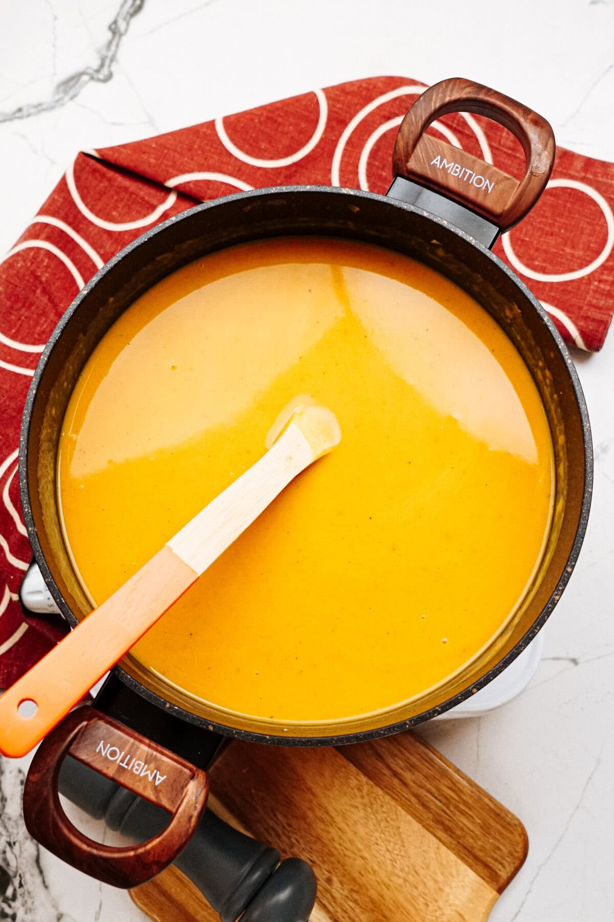 A pot of smooth, orange soup with a wooden spoon, placed on a wooden cutting board and a red cloth. The pot has "AMBITION" engraved on the handles.