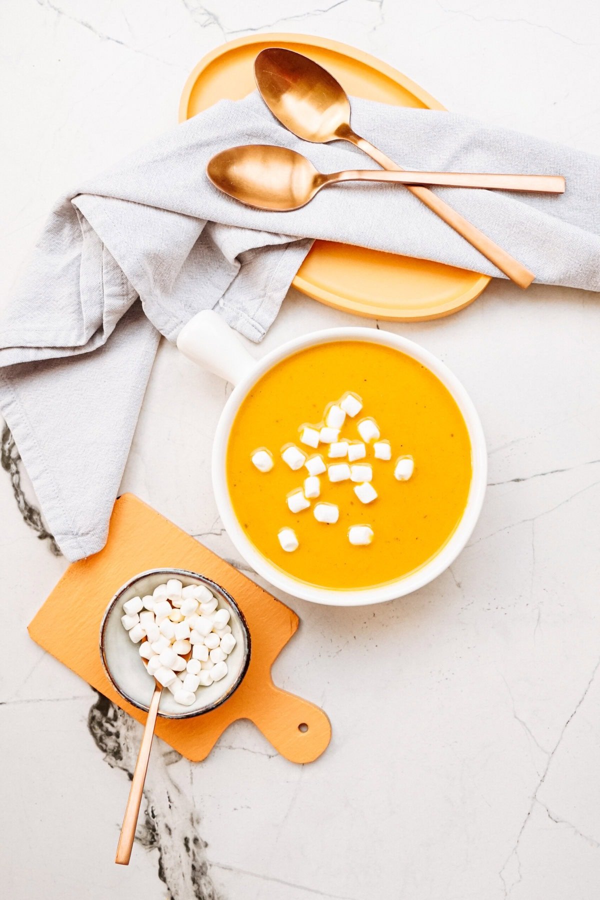 A bowl of curry soup topped with marshmallows sits on a marble surface, accompanied by a dish of marshmallows, two brass spoons, and a folded napkin.