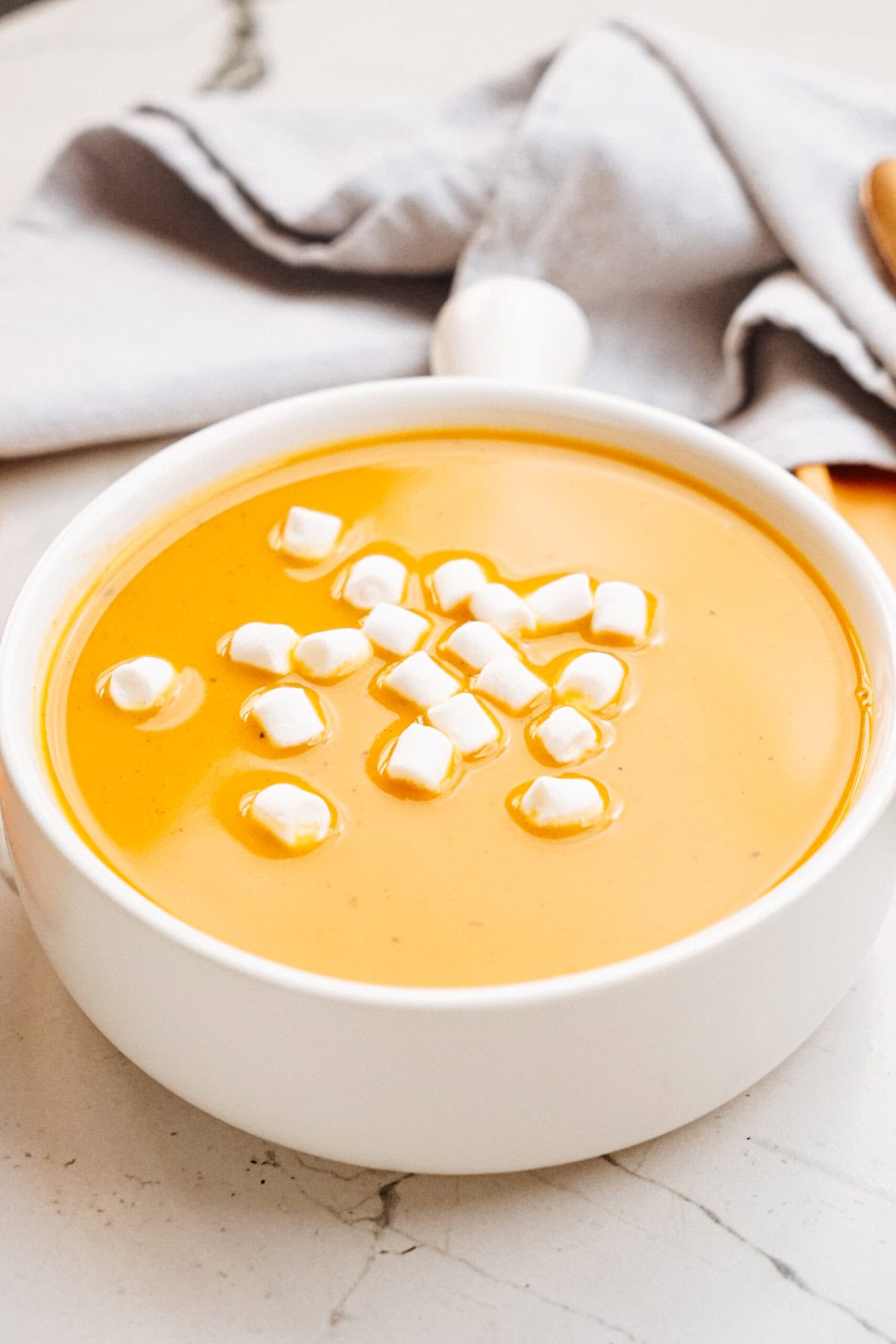 A white bowl filled with orange soup, topped with small white marshmallows. A gray cloth napkin is in the background.