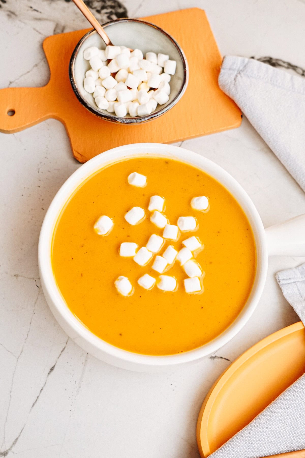 A bowl of creamy orange soup topped with small marshmallows, placed next to a small bowl of additional marshmallows on a cutting board, and a folded cloth napkin.