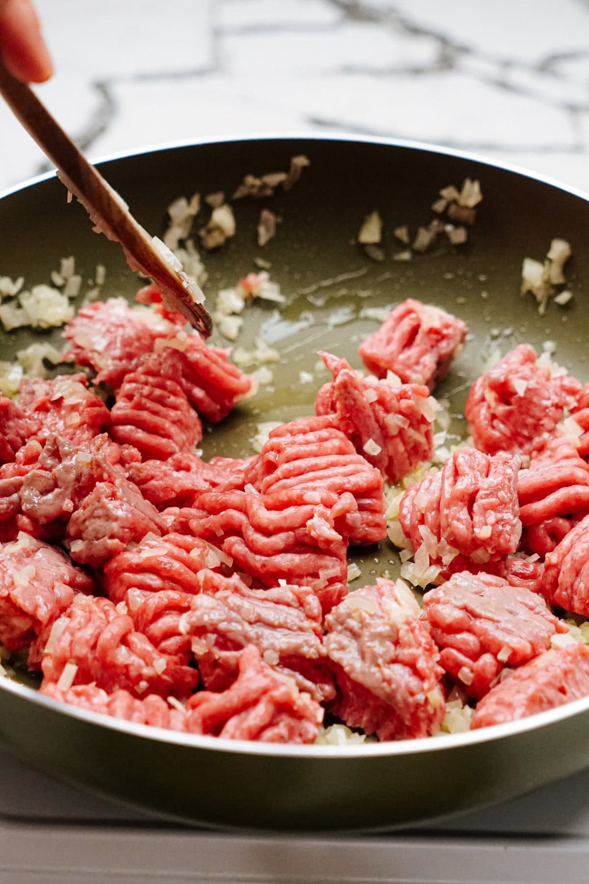 A frying pan contains raw ground beef and chopped onions being stirred with a wooden spatula on a stove.