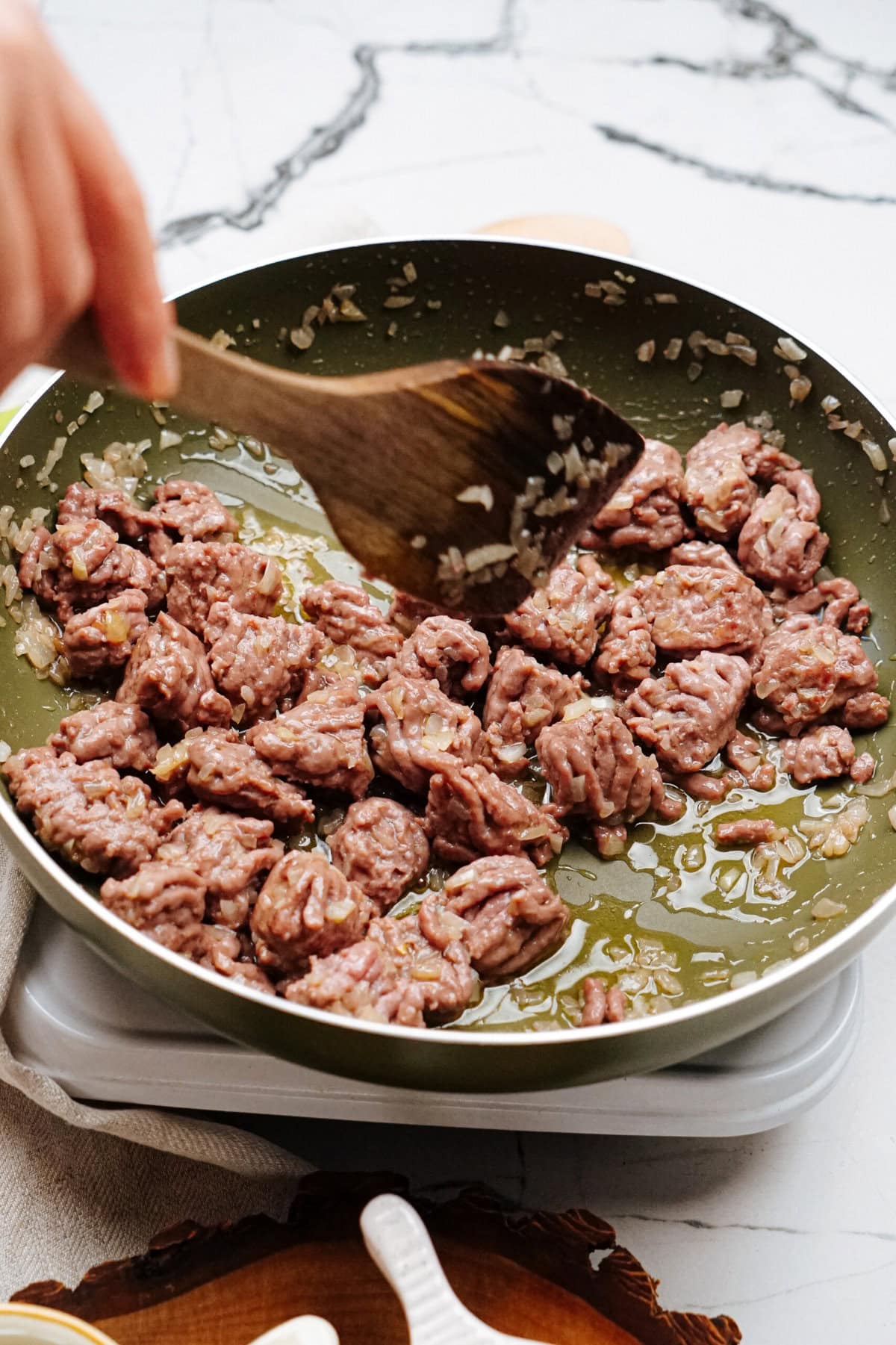 A person stirs ground beef with onions cooking in a green frying pan on a stove.