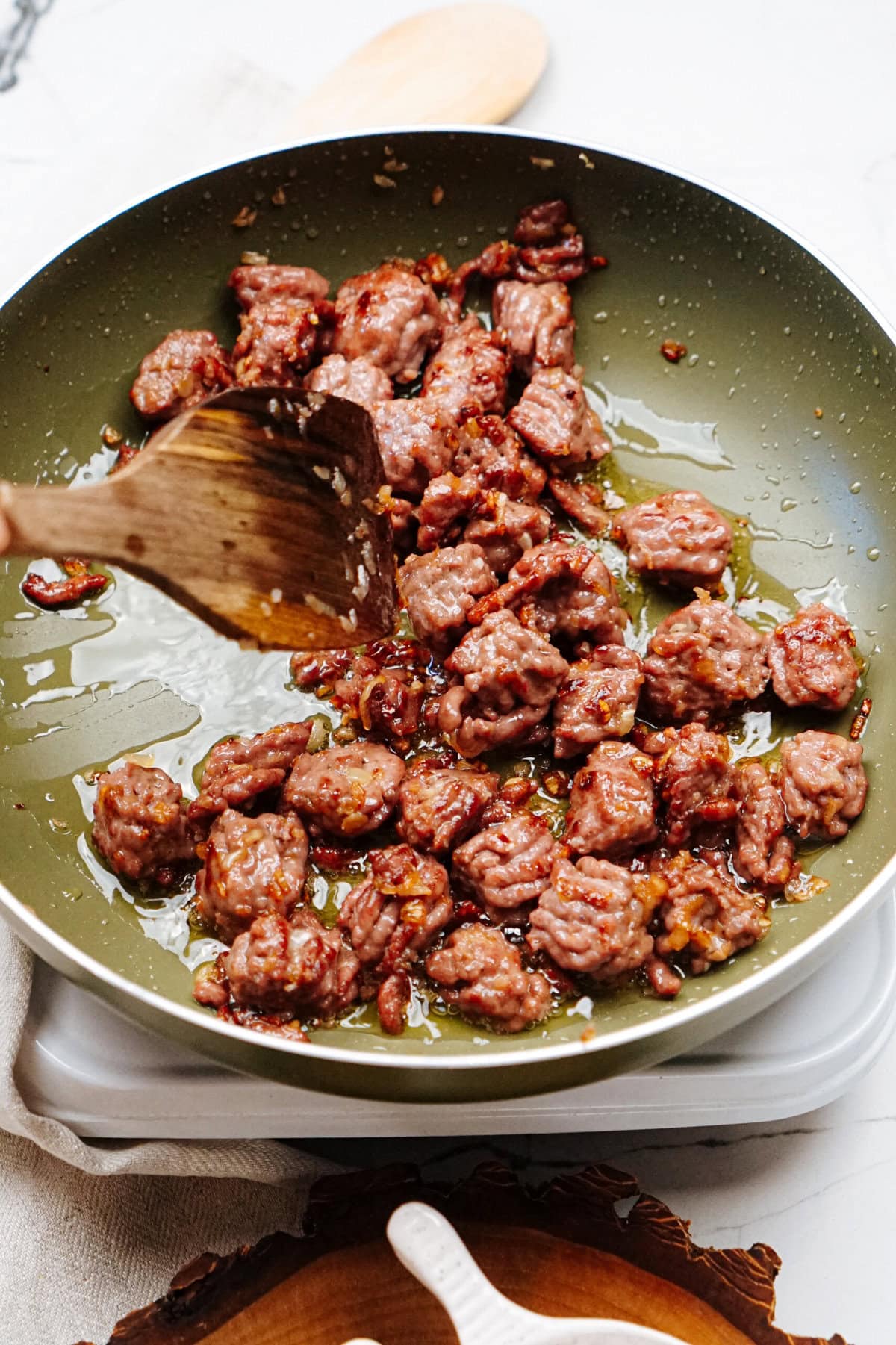 Ground meat pieces being cooked in a green non-stick pan, stirred with a wooden spatula.