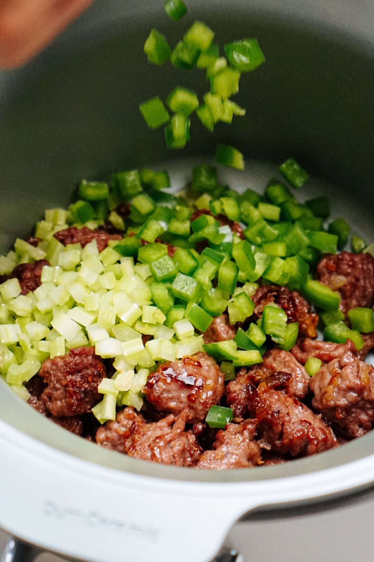 A pot with browned ground meat, chopped celery, and green bell peppers being added on top, ready for cooking.