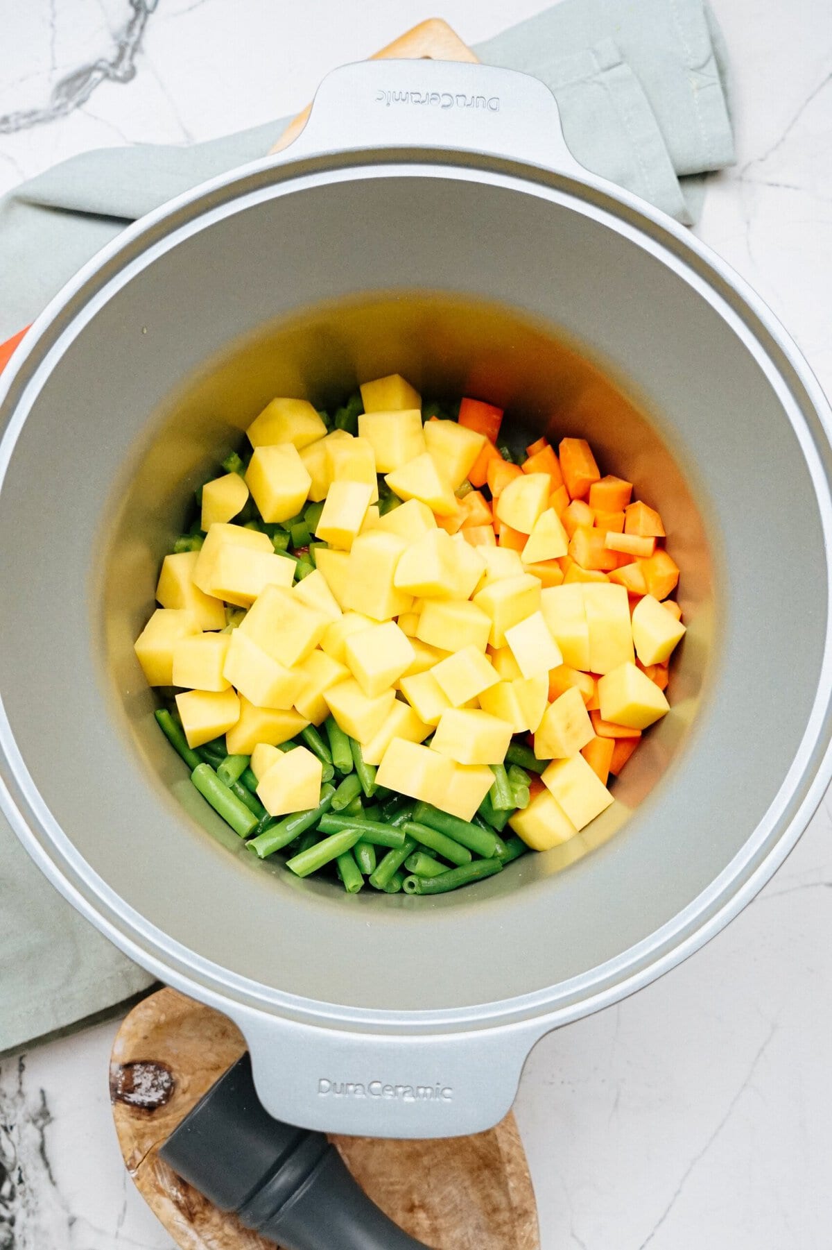 A pot filled with chopped vegetables, including yellow potatoes, green beans, and orange carrots, ready for cooking. The pot is placed on a wooden cutting board with a folded kitchen towel underneath.