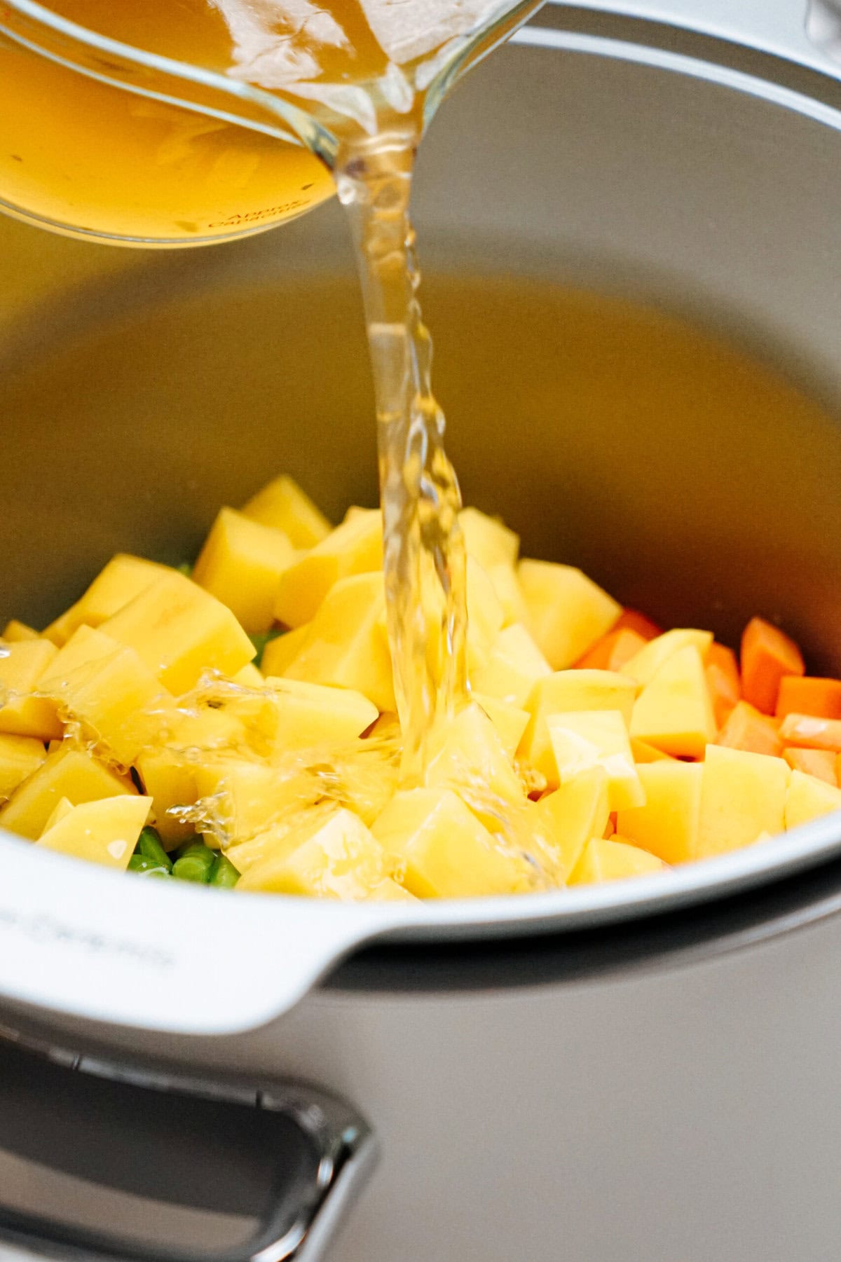 A pot with chopped potatoes, carrots, and celery, being filled with broth poured from a glass container.