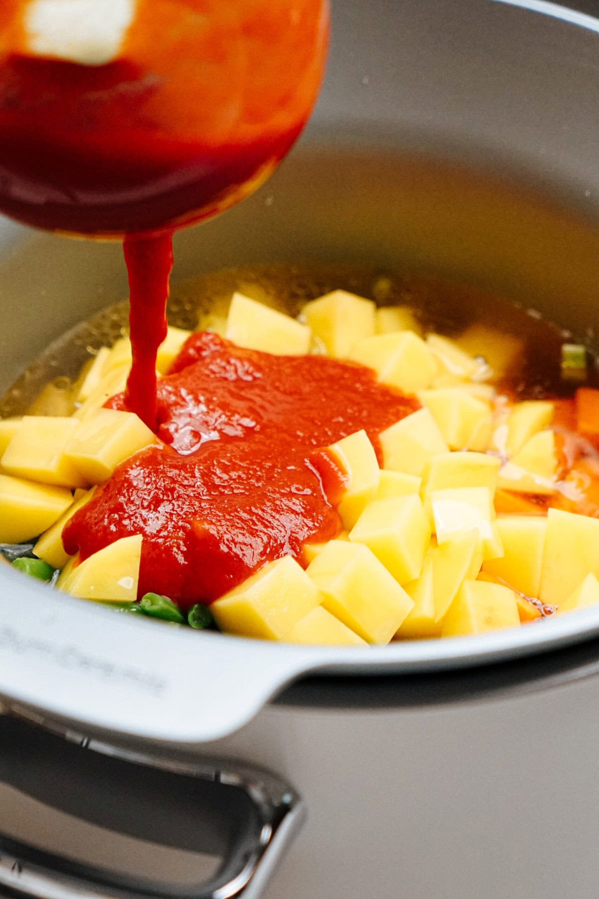 A pot filled with cubed yellow vegetables and diced green vegetables, with tomato sauce being poured over them.