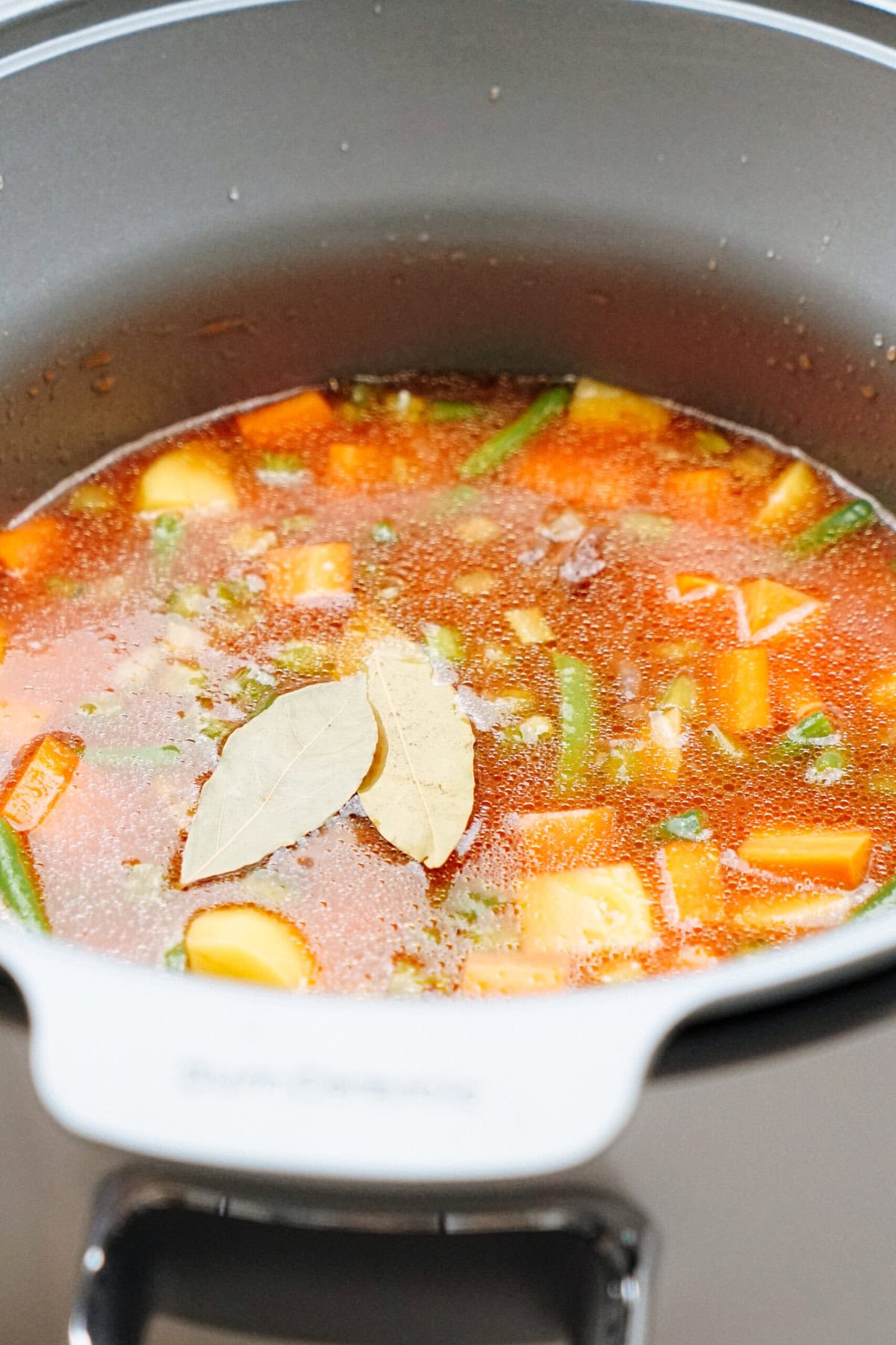 A pot of vegetable soup with visible chunks of carrots, potatoes, green beans, and two bay leaves floating on the surface.