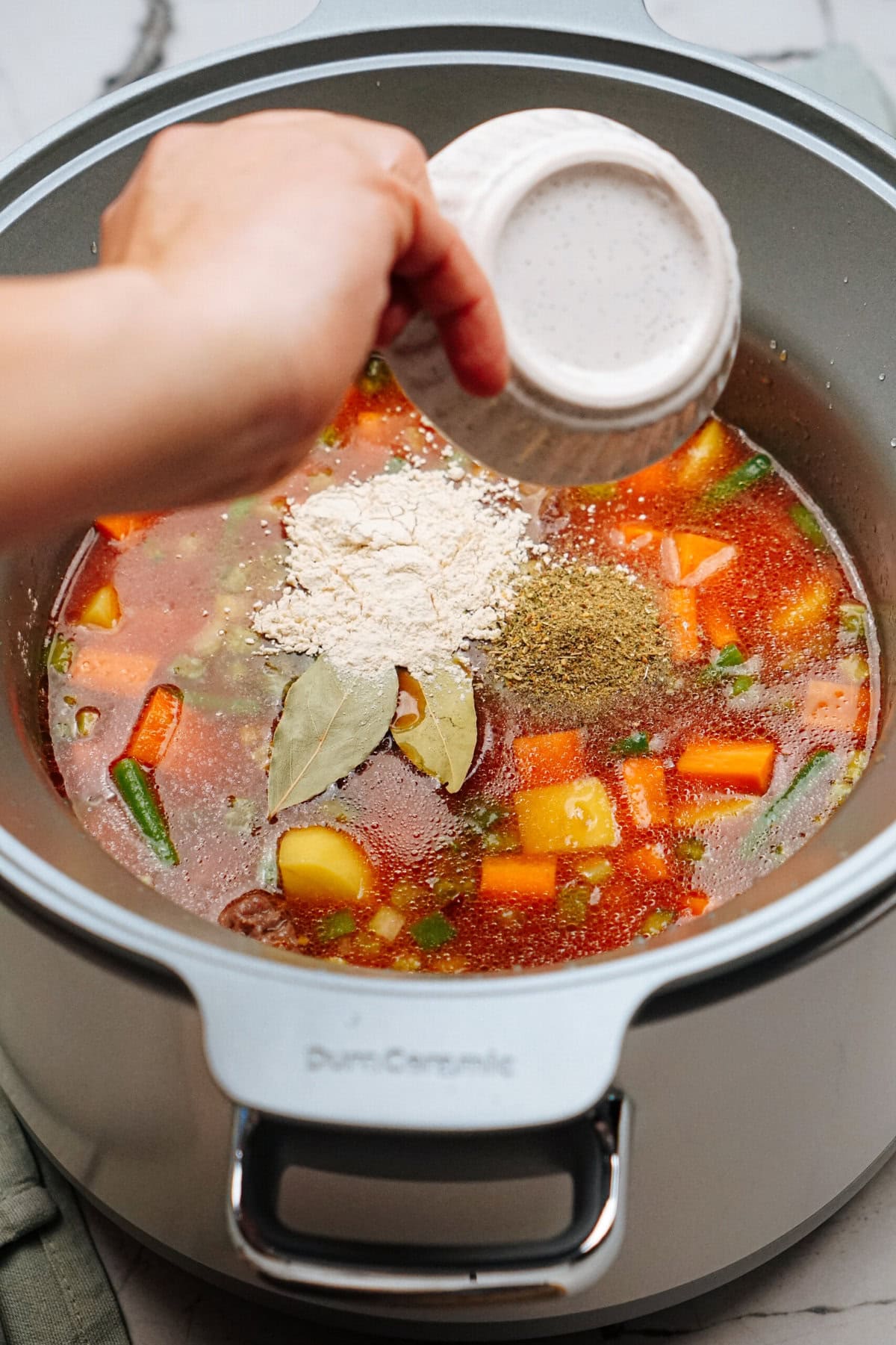 A hand is adding flour and spices into a pot filled with a colorful vegetable stew. The pot contains carrots, green beans, potatoes, and a rich broth.