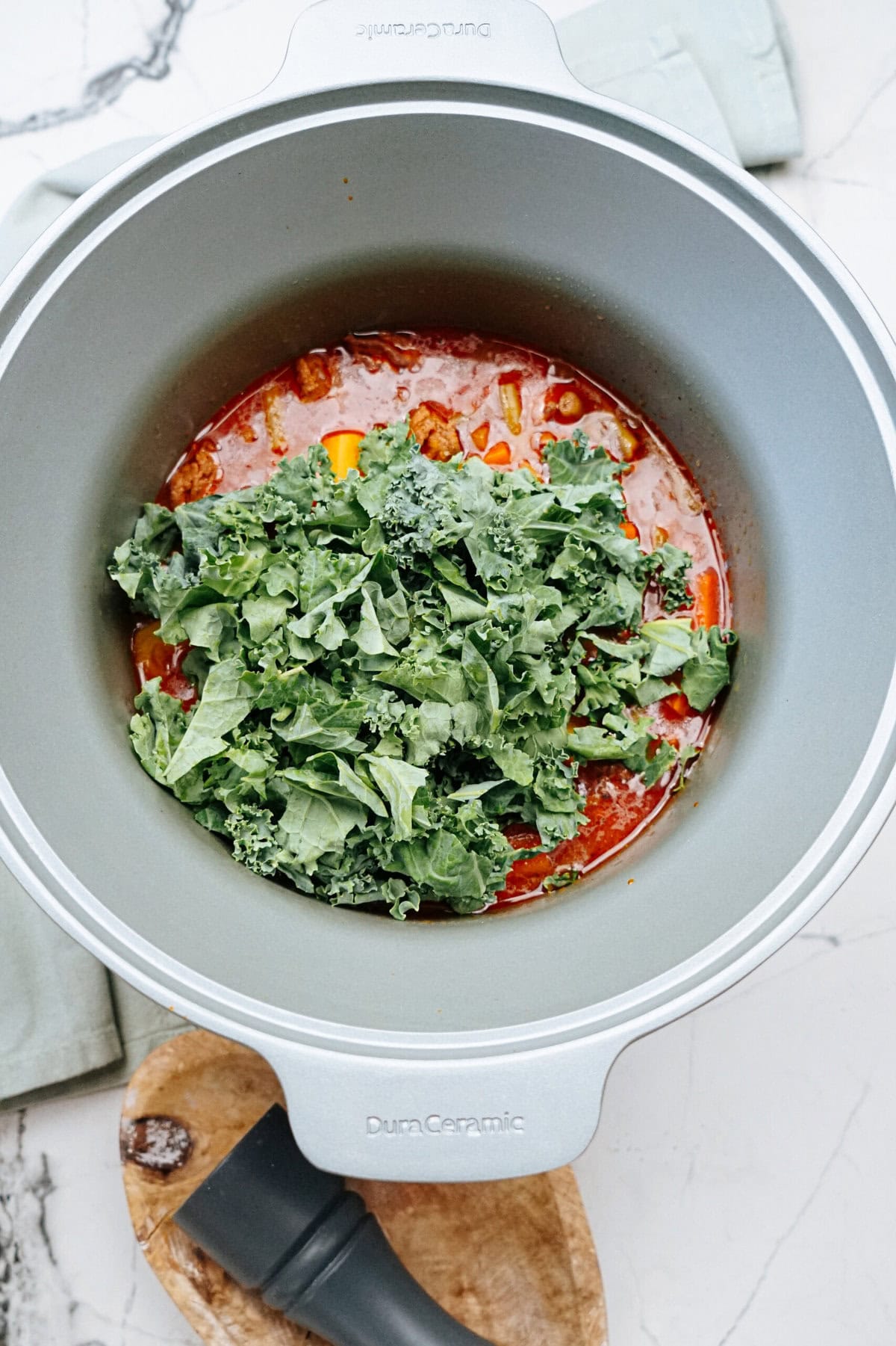 A pot filled with a tomato-based stew and topped with fresh green kale. The pot is placed on a white marble countertop with a wooden spoon beside it.