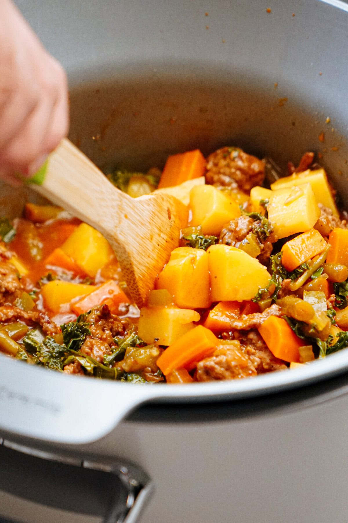 A person stirs a pot of chunky vegetable stew using a wooden spatula. The stew contains various vegetables including carrots, potatoes, and greens.