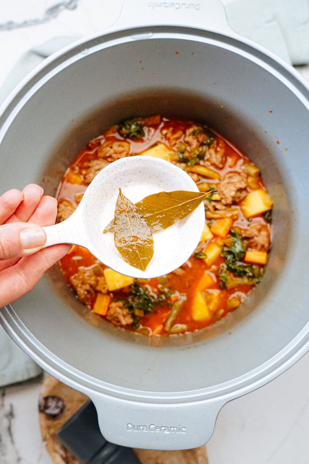 Hand adding bay leaves to a pot of soup with meat, potatoes, and leafy greens.