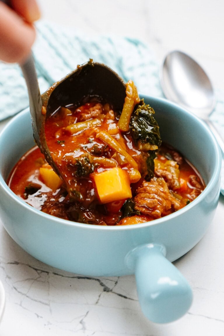 A ladle serves a portion of hearty vegetable and meat stew into a blue bowl on a marble countertop, with a spoon and blue napkin in the background.