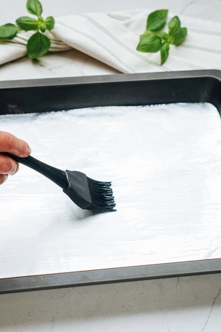 Hand using a black brush to coat a baking sheet with a white substance. Fresh basil leaves and a striped cloth are in the background.